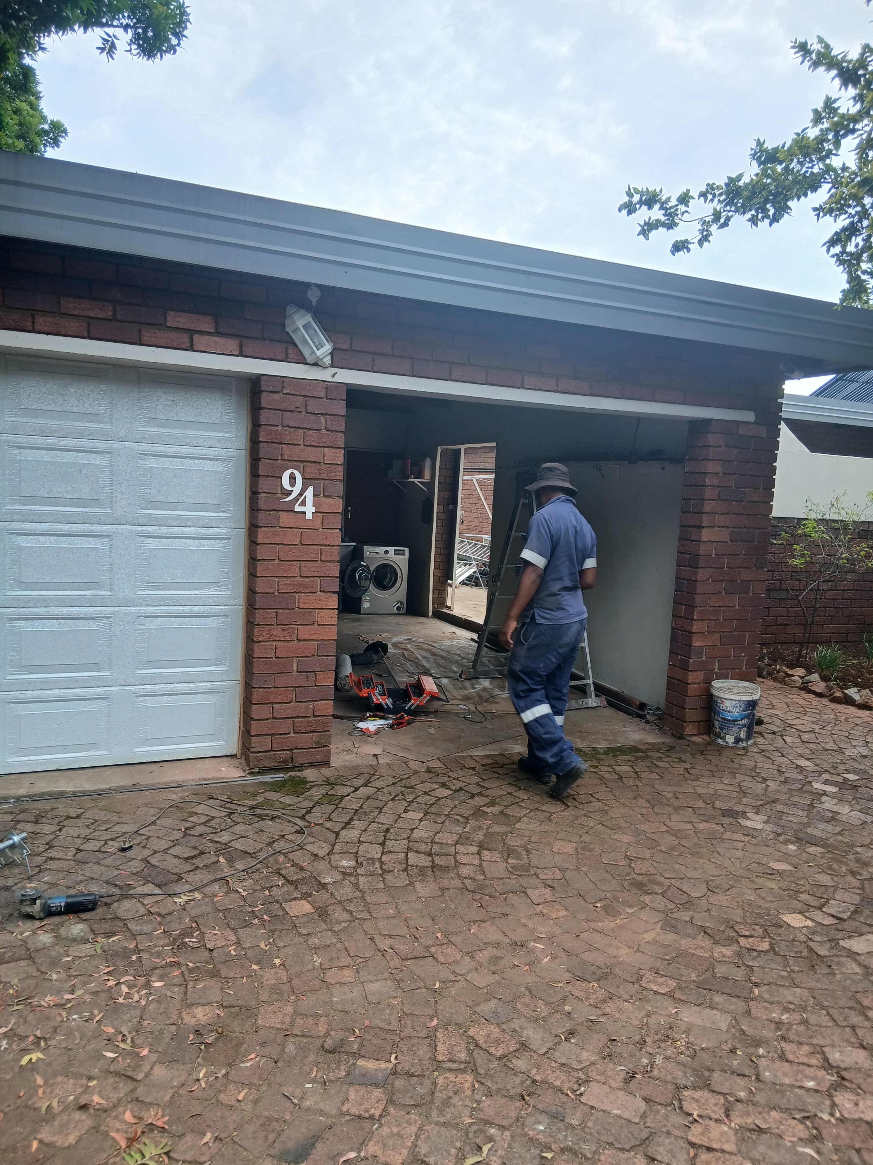 Man in blue workwear enters a brick garage with tools on a brick driveway.
