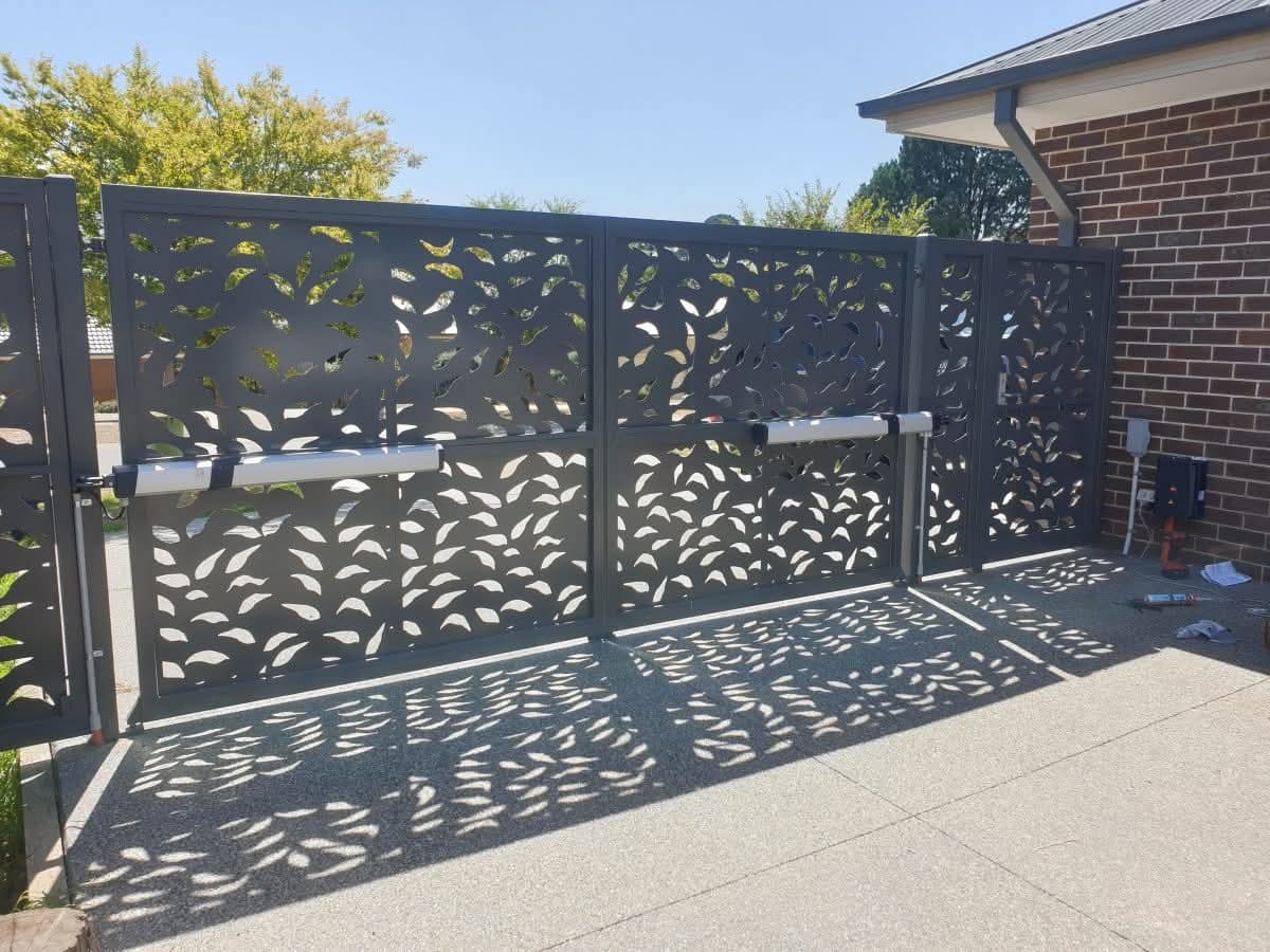 Decorative dark grey metal gate with leaf-shaped cutouts casting shadows on a sunny driveway.