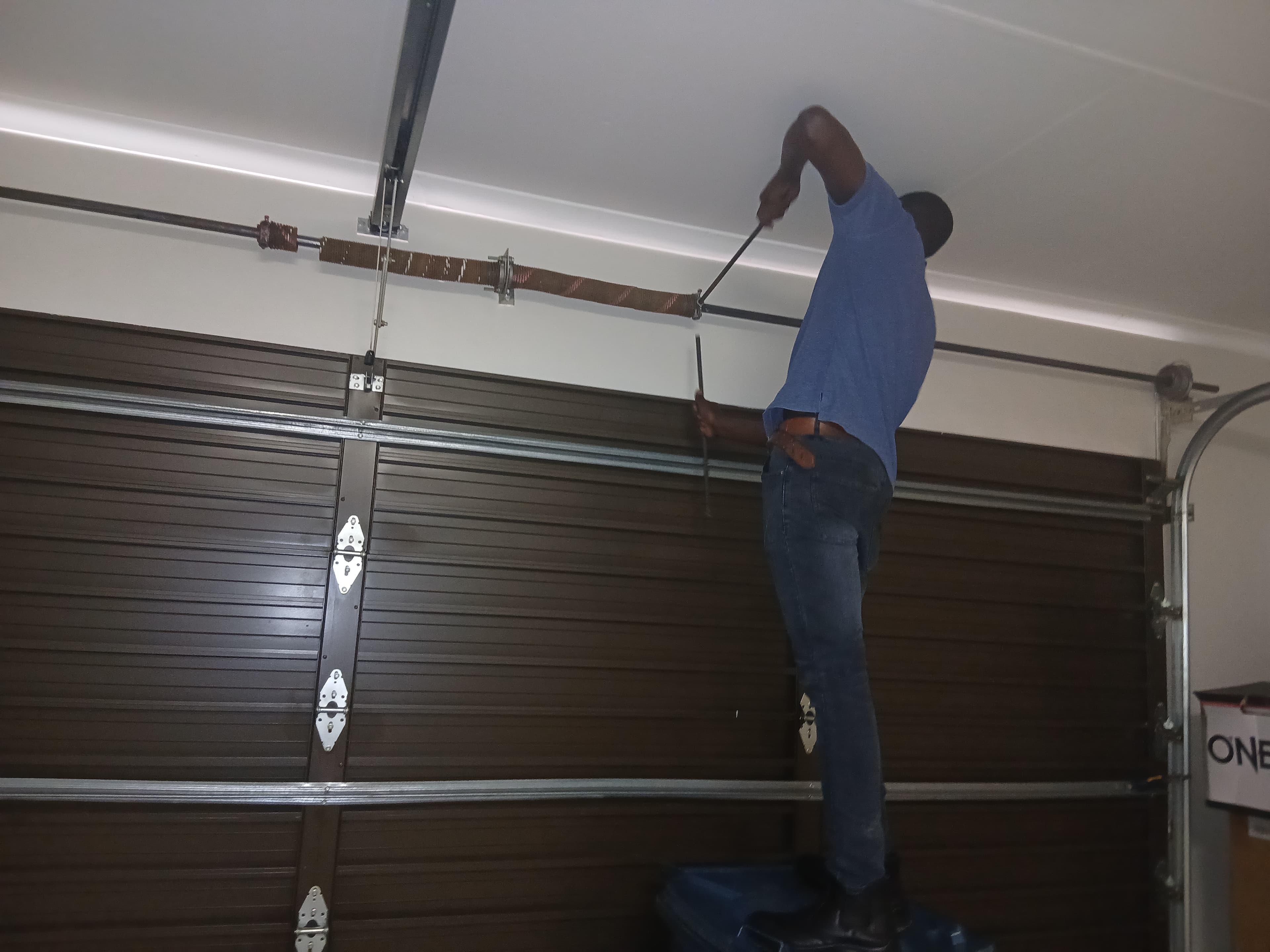 Technician in blue shirt repairing a brown garage door torsion spring using metal winding bars.