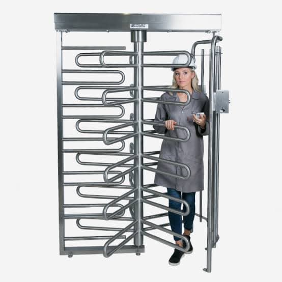 Woman wearing a hard hat and lab coat passing through a full-height metal turnstile.