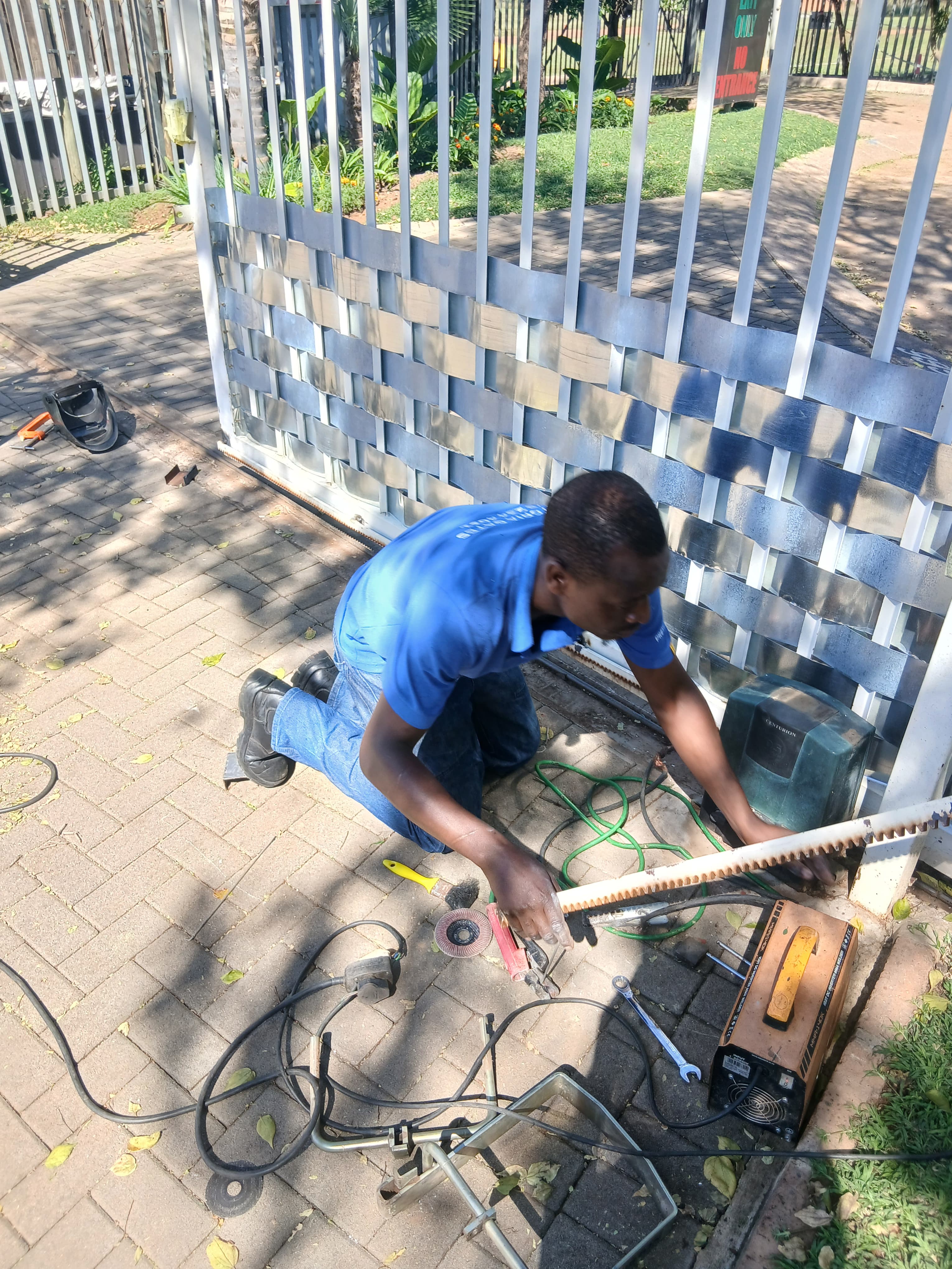 Man in blue shirt repairs a metal gate mechanism on a brick patio with tools.