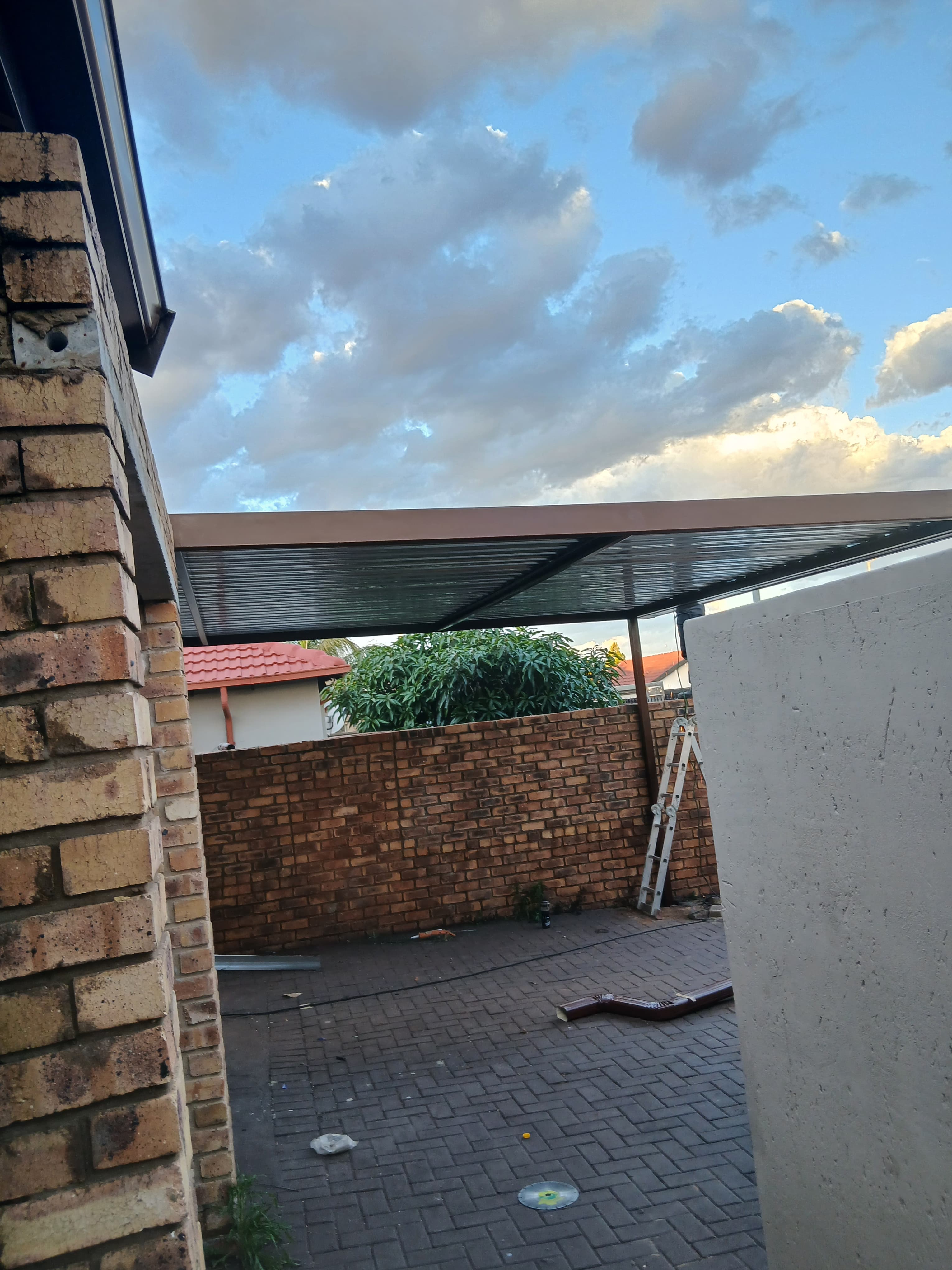 Metal carport structure over a brick-walled patio with a ladder under a cloudy sky.