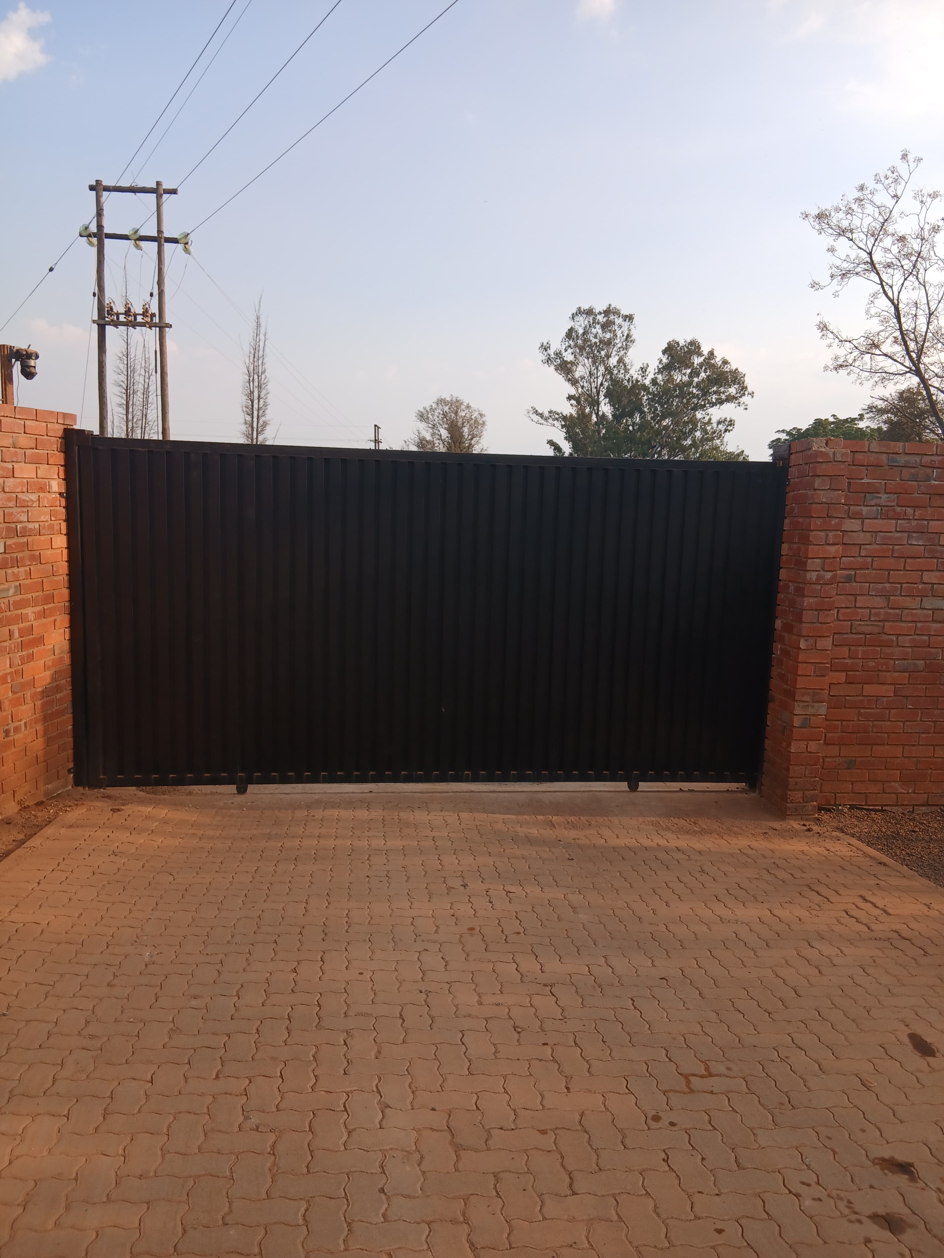 Large black metal gate between red brick pillars on a patterned brick paved driveway.