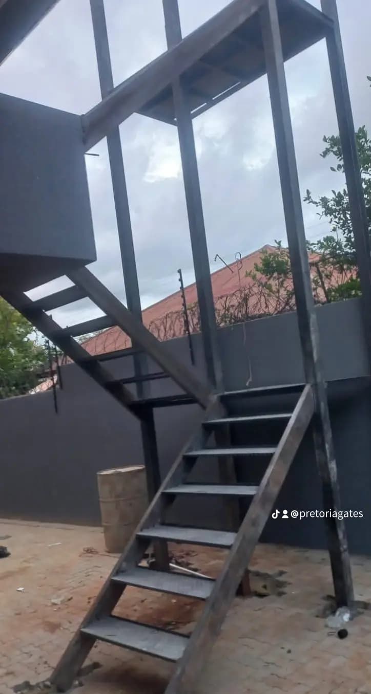 Dark metal outdoor staircase with two flights and a landing against a grey wall.