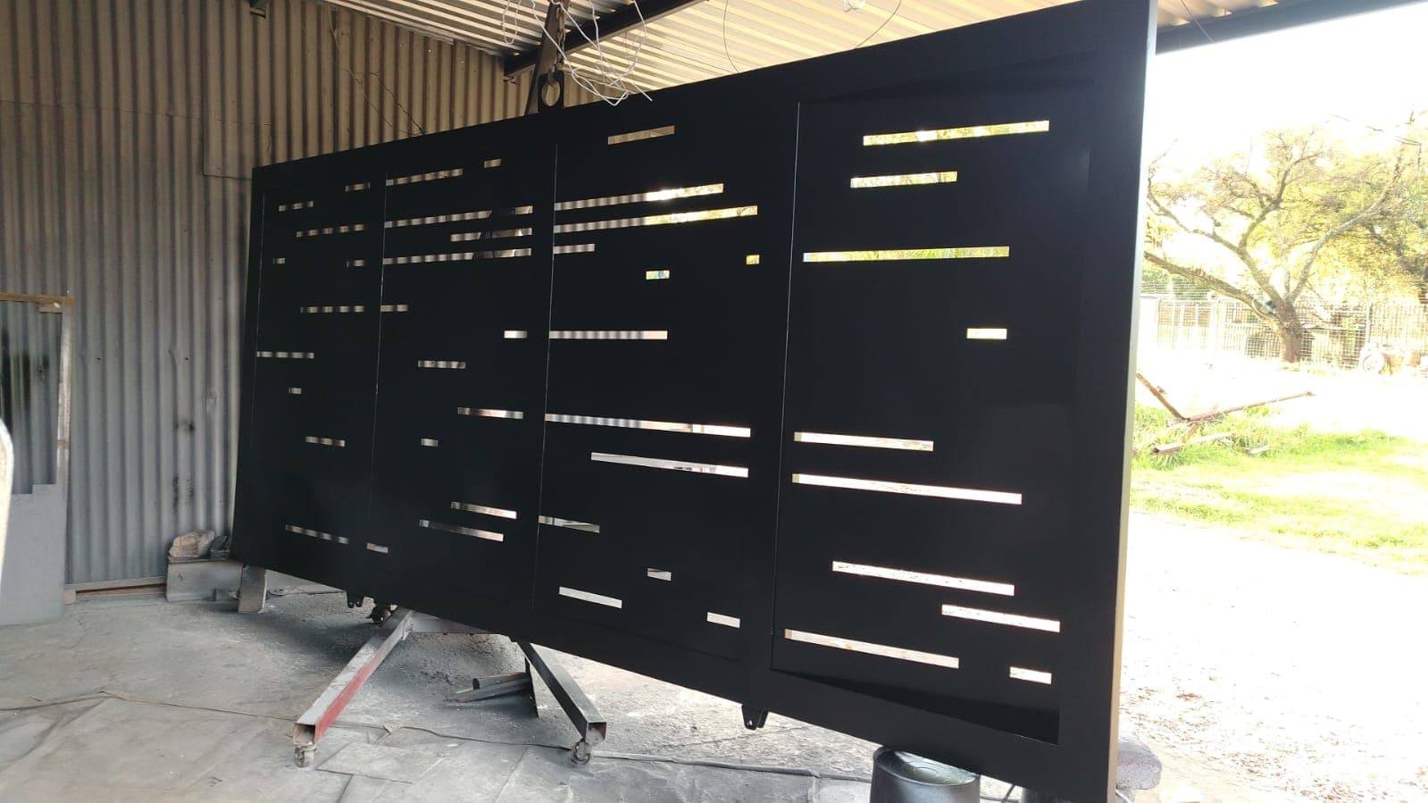 Modern black metal gate with decorative horizontal laser-cut slits, standing in a fabrication workshop.