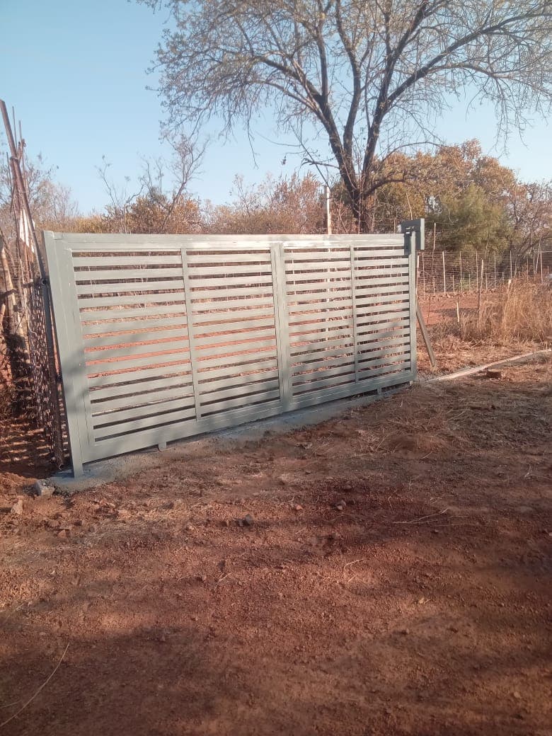 Grey horizontal slatted metal gate on red dirt in a rural landscape with sparse trees.