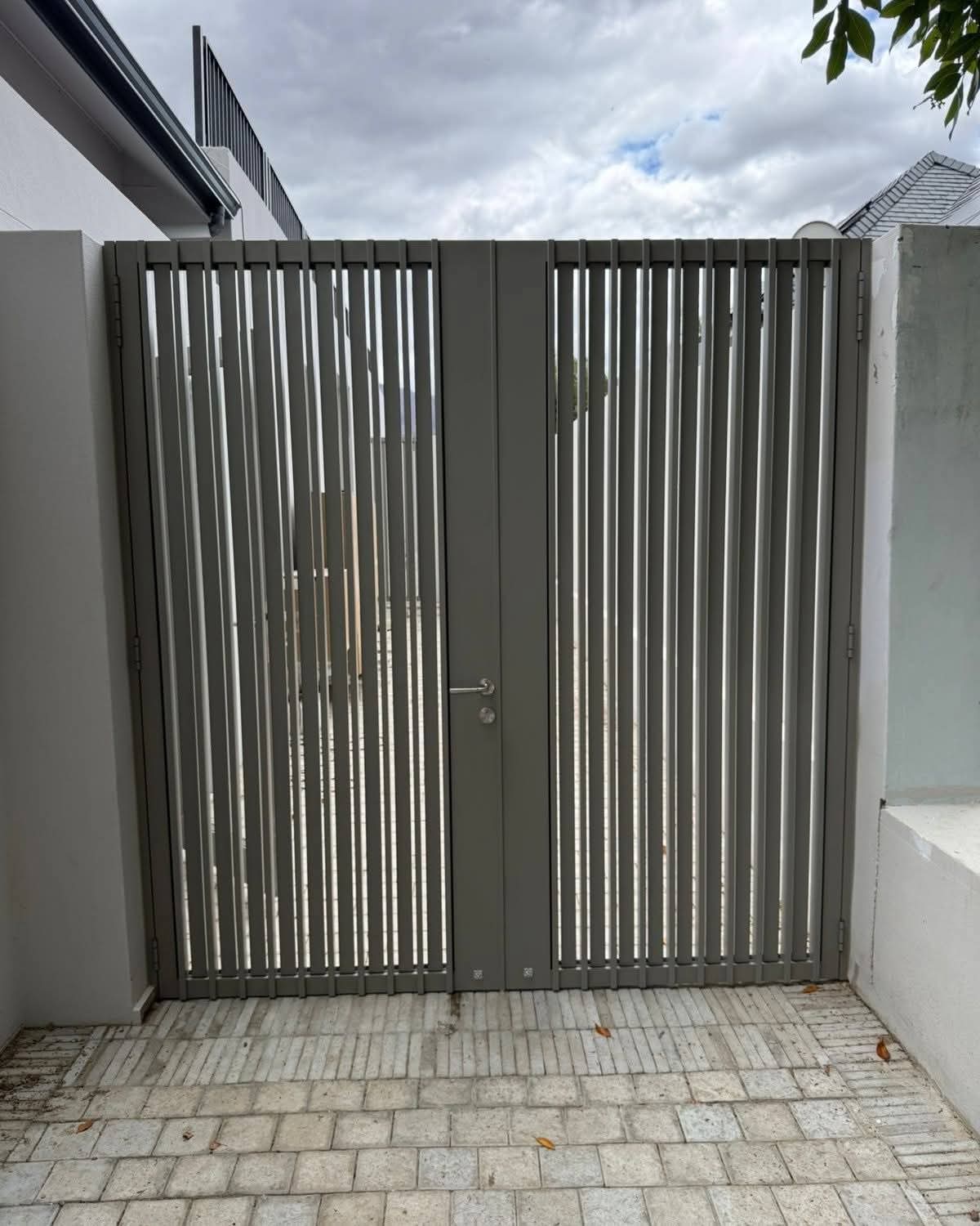 Modern grey vertical slat gate between white walls on a light stone paved path.