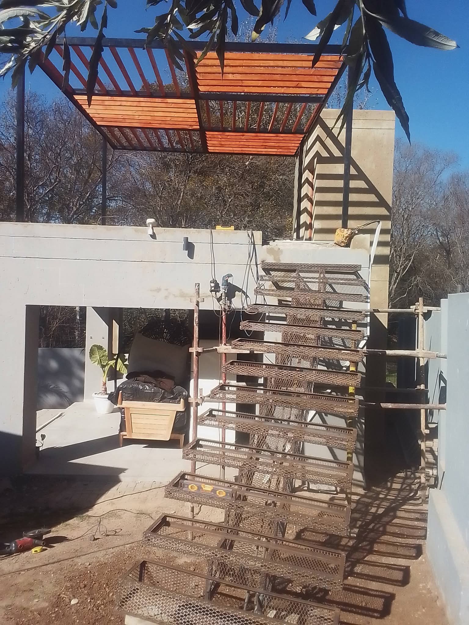 Metal mesh staircase under construction next to a concrete building with a wooden slat pergola.