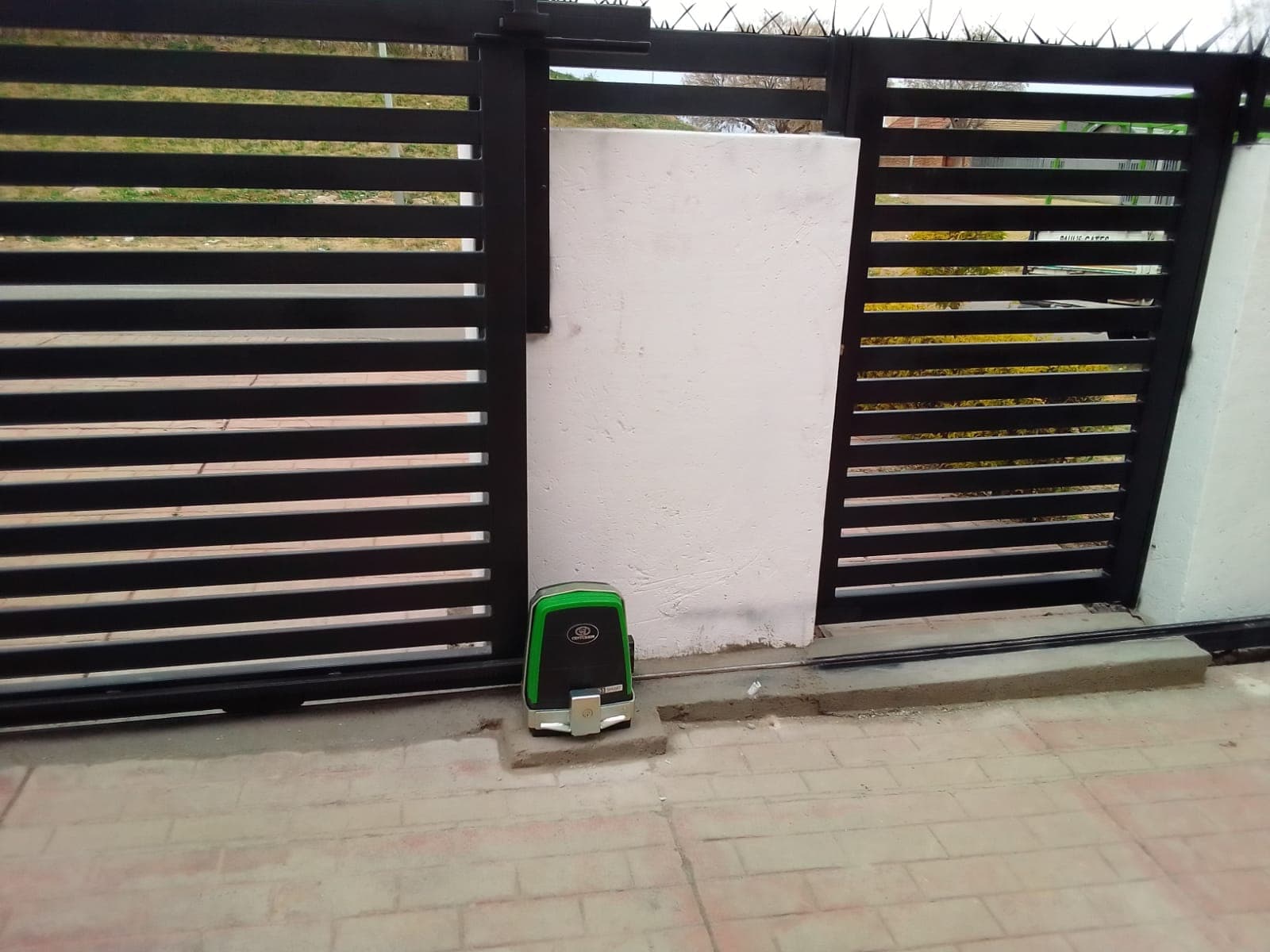 Black horizontal slat sliding gate with a green automatic motor and white concrete pillar.