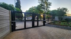 Modern black metal double swing gate with automatic openers on a brick paved residential driveway.