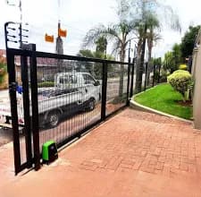 Black sliding mesh gate with green motor and electric fencing on a brick driveway.
