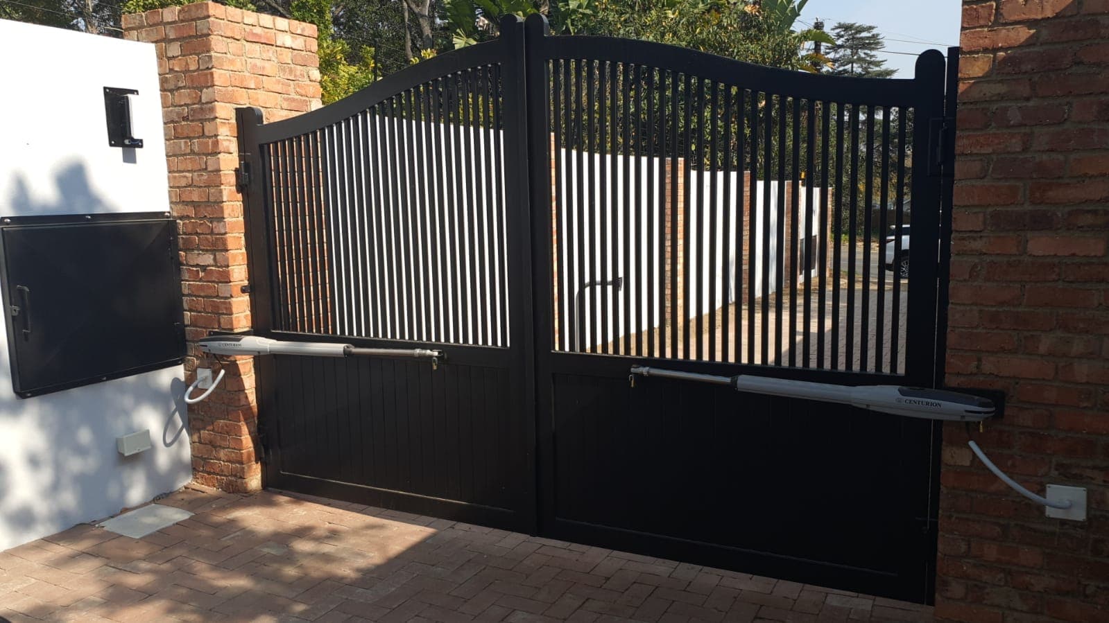 Black double-swing automatic driveway gate with vertical bars and solid panels between brick pillars.