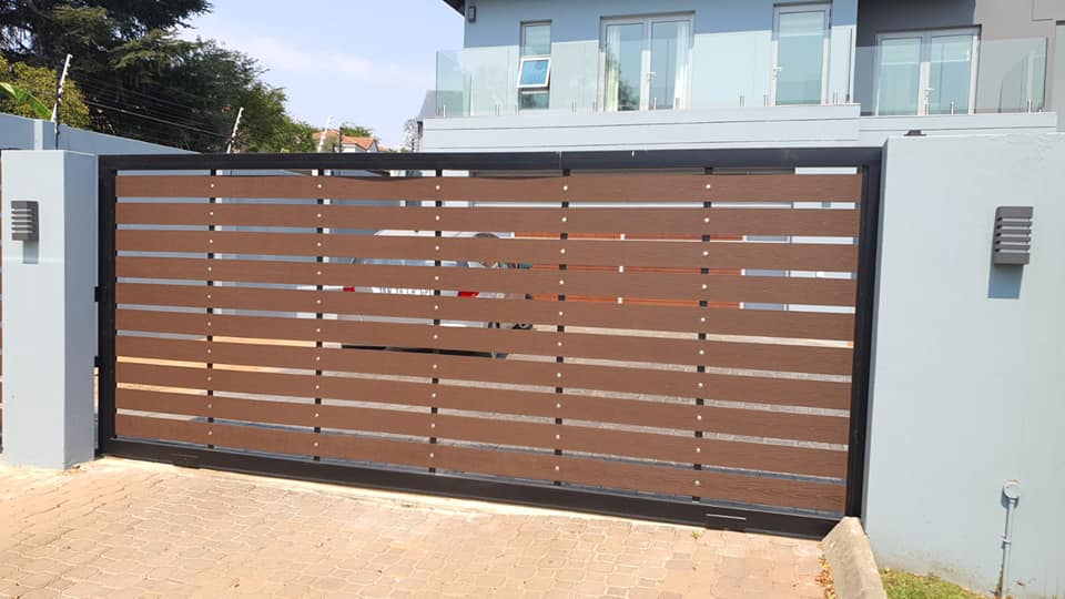 Modern driveway gate with horizontal brown slats and black frame between two grey pillars.