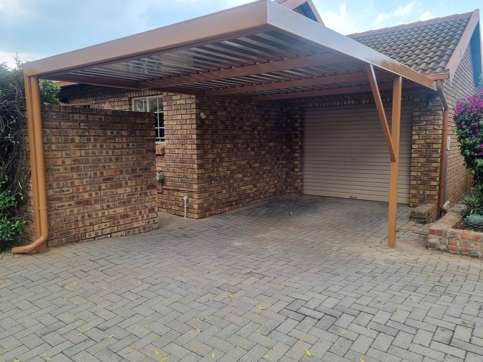 Brown metal carport attached to a brick house over a paved herringbone driveway.